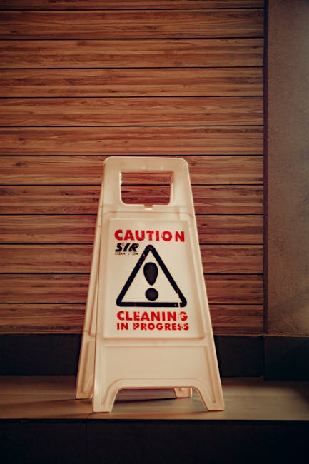 A person wearing blue disposable gloves is using a blue microfiber cloth to clean the surface of a black remote control, which rests on a white table or countertop. The background appears to be a soft
